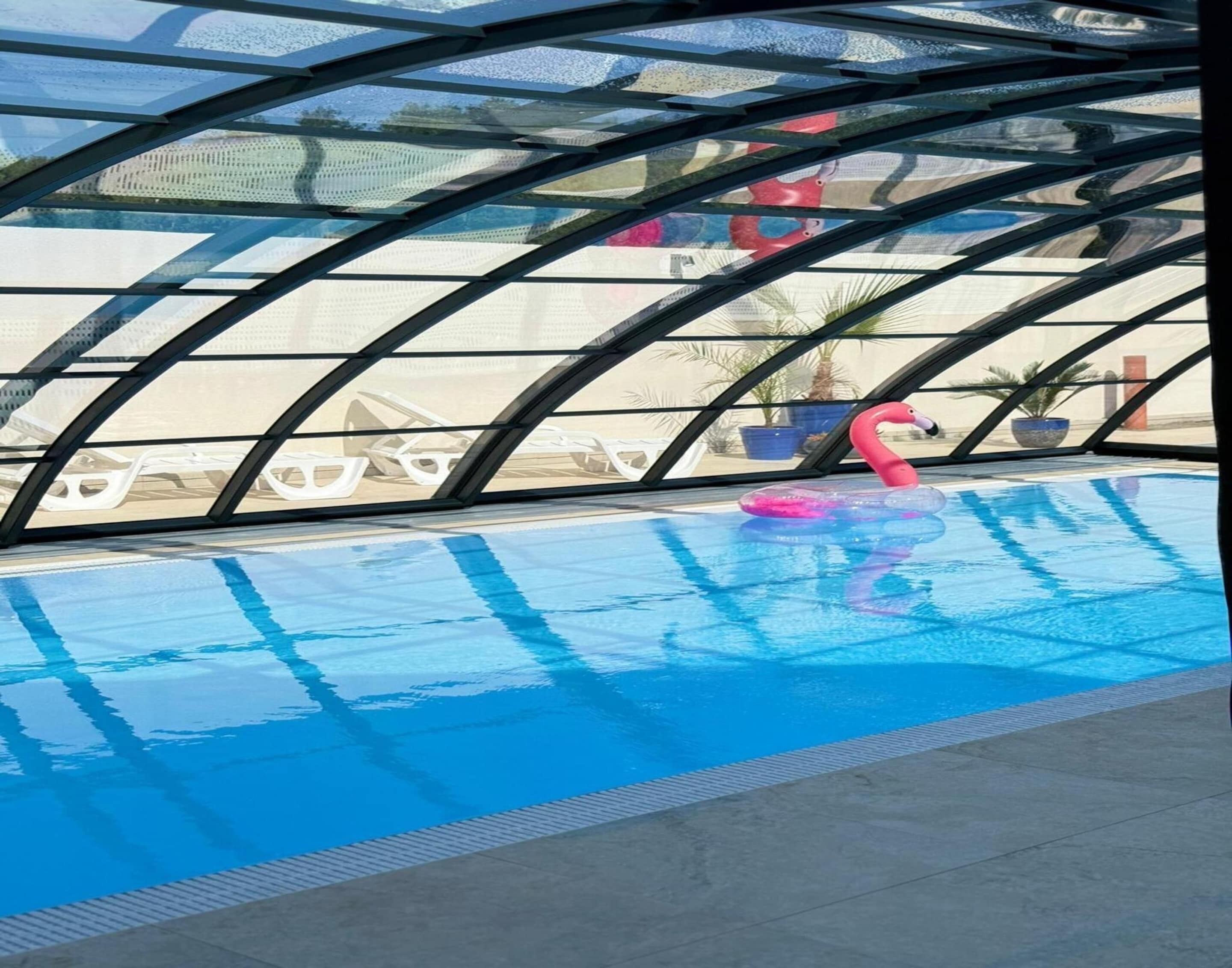 En inomhuspool med kristallklart blått vatten, en rosa uppblåsbar flamingofigur och solstolar under ett välvt glastak.
