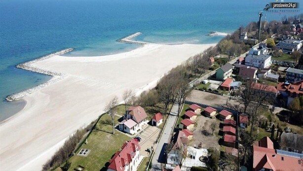Una vista aerea di una zona costiera con spiaggia sabbiosa, edifici residenziali e un molo che si protende verso il mare.