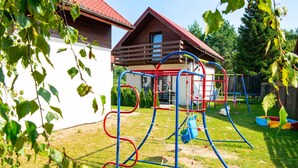 Un'area giochi con altalene e strutture per arrampicarsi, circondata da una casetta di legno e un muro bianco.