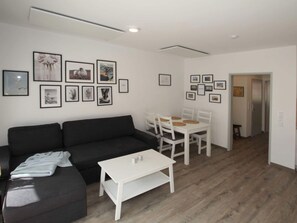 A modern living room with a black sofa, a white coffee table, and a dining area with a white table and chairs. The walls are adorned with framed pictures. - Ferienwohnung in Ruhiger Lage in Dornumersiel (Dornum)