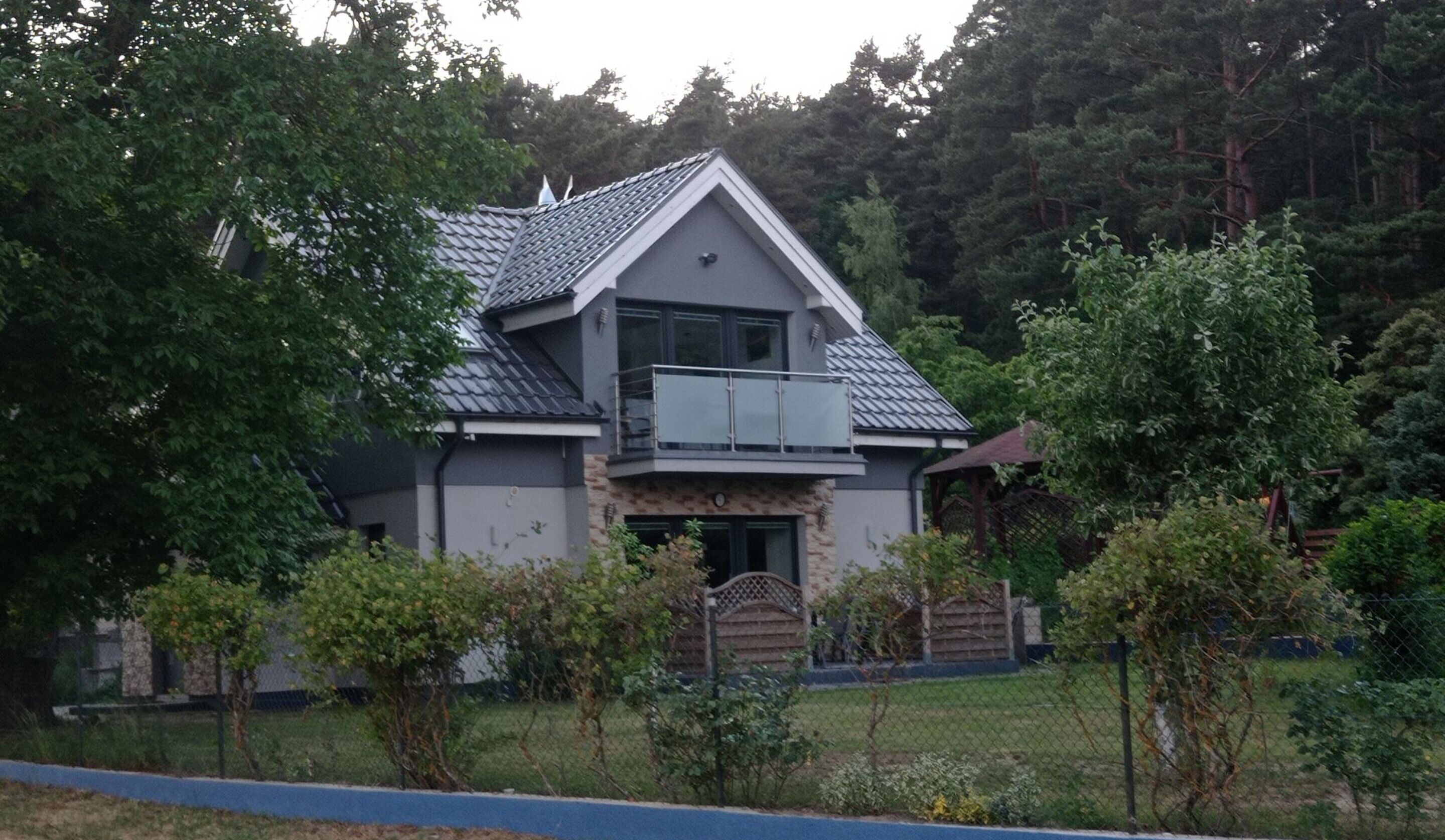 A modern two-story house with a balcony, surrounded by lush greenery and trees.
