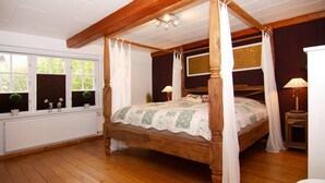 A four-poster canopy bed with a wooden frame, white curtains, and a floral bedspread. There is a window with a view of greenery outside, a bedside table with a lamp, and a radiator under the window. - Ferienhaus in Rehm-flehde-bargen mit Gemeinsamer Sauna (Rehm-Flehde-Bargen)
