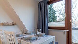 A dining area with a white table set for four, a window with blinds, and a view of trees outside. - Comfortable Holiday Apartment in St. Peter Ording (St. Peter-Ording)