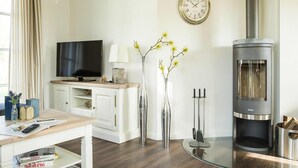A living room with a TV, a fireplace, a clock, and a vase with flowers. - Reetdachhaus am Bodden mit Sauna (Fuhlendorf)