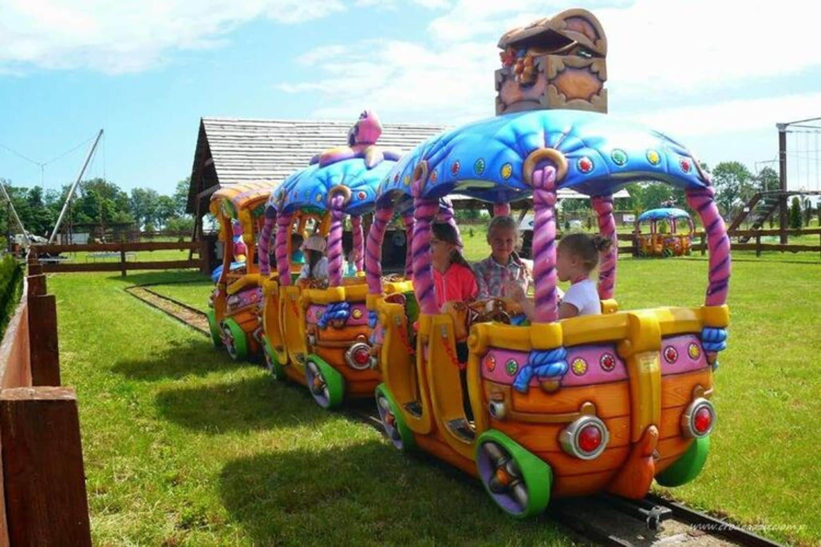 A colorful train ride with passengers, set on a grassy track with a wooden fence and a building in the background.