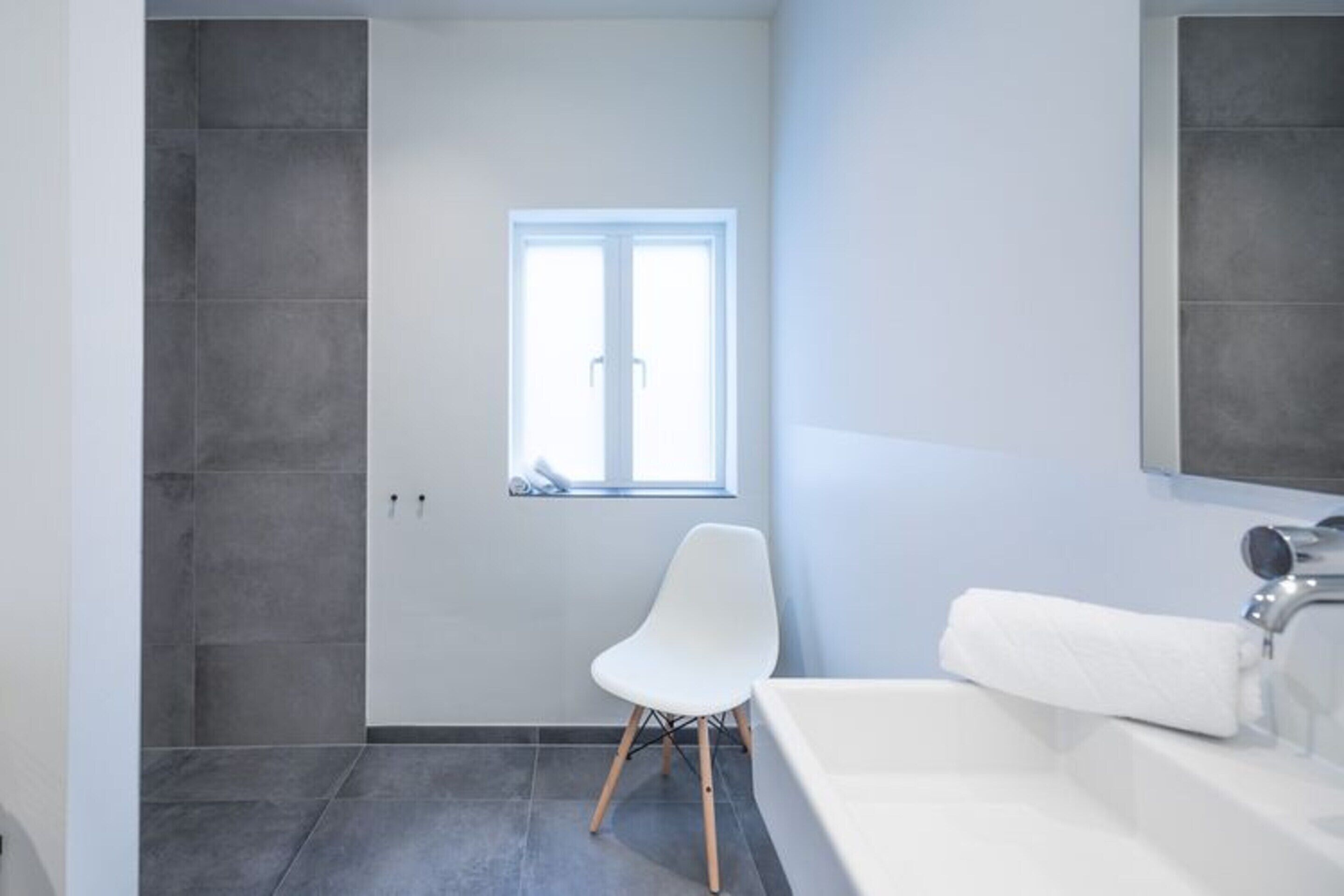 A modern bathroom with a white bathtub, a white chair, and a window providing natural light.