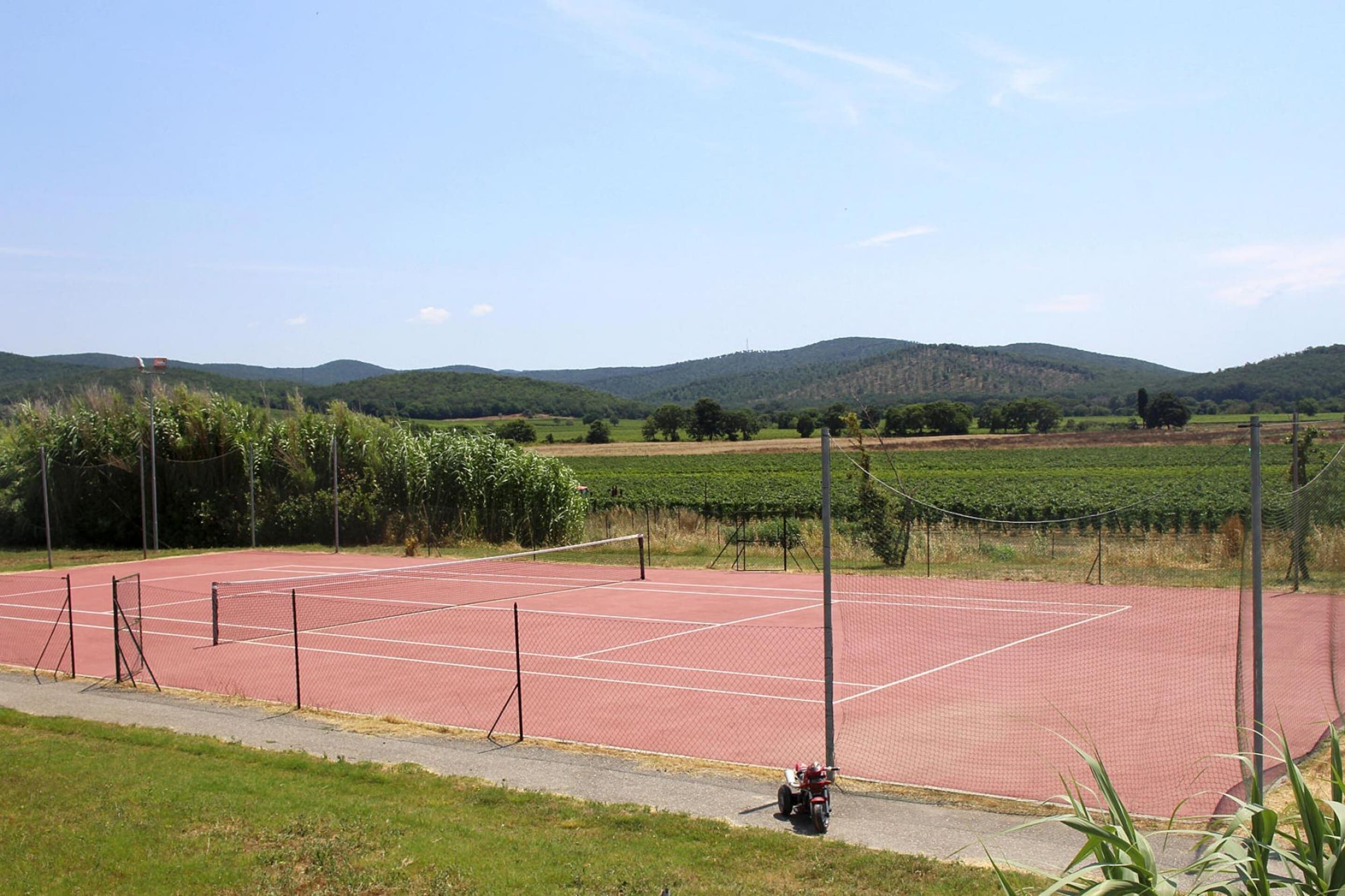Tenniškenttä, jossa on verkko, ja jonka ympärillä on aita. Kentältä lähtee sorapolku pysäköityyn ajoneuvoon.