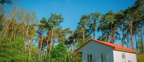 A small, single-story house with a red roof, surrounded by a lush green landscape and tall trees.