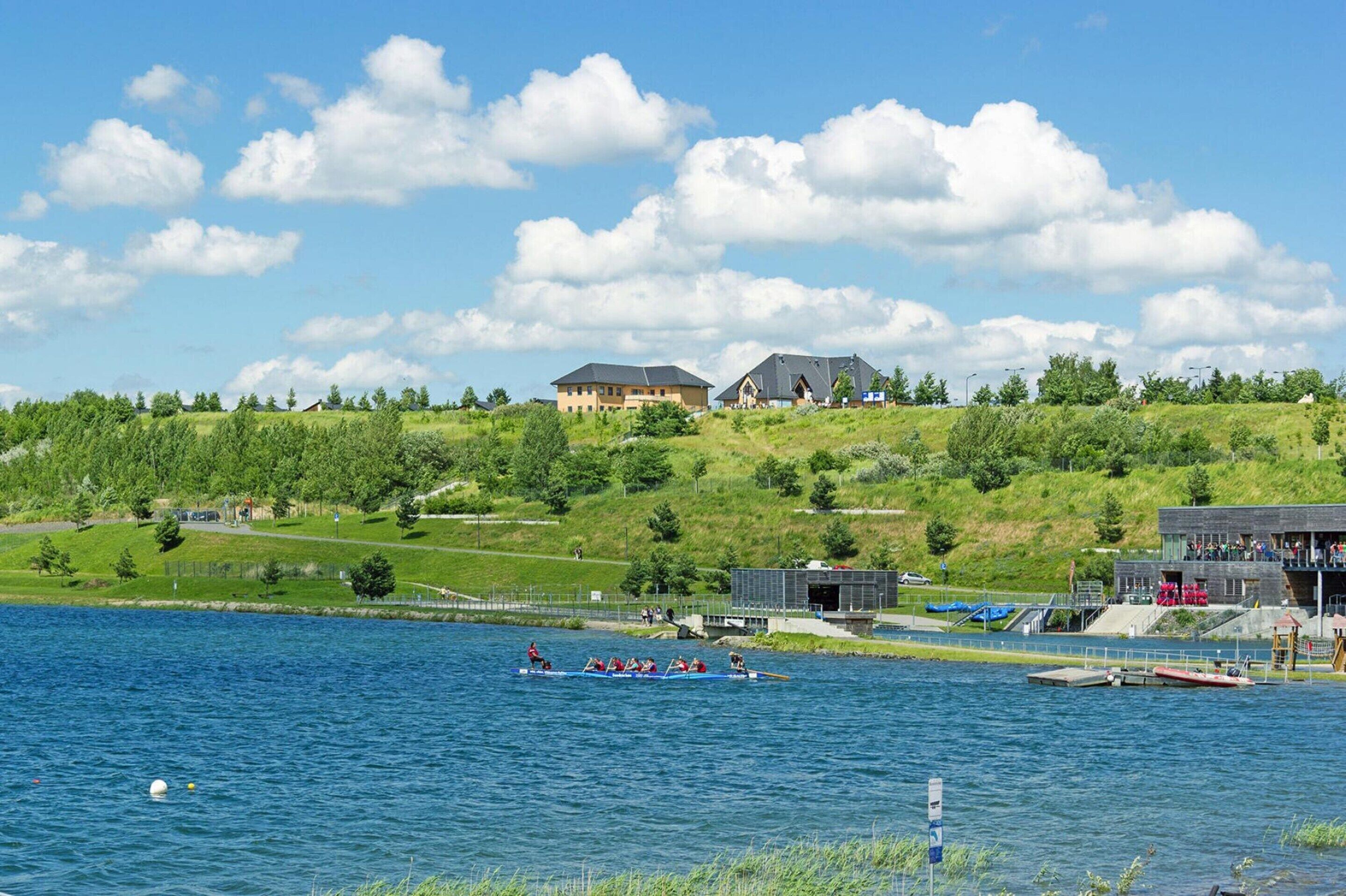 A lakeside view with a rowing team in a boat, a building with a sloped roof, and a grassy hill with trees.