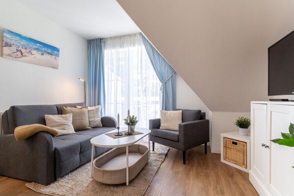 A modern living room with a sofa, armchair, and coffee table. There is a television on the right and a painting on the wall. - Ferienwohnung in St. Peter-ording (St. Peter-Ording)