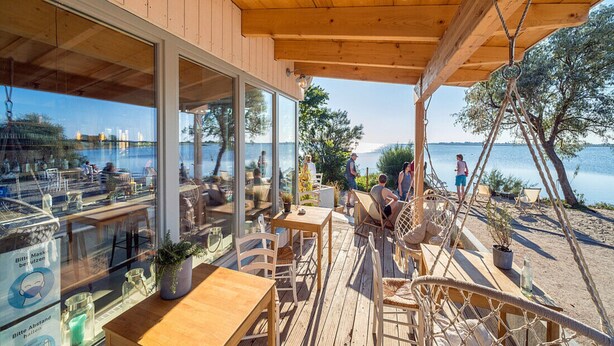A wooden deck with outdoor seating, large windows overlooking a body of water, and a view of the surrounding landscape.