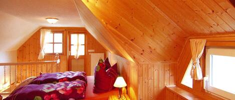 A cozy attic bedroom with a wooden sloped ceiling, a bed with colorful bedding, a bedside lamp, and a window with curtains.