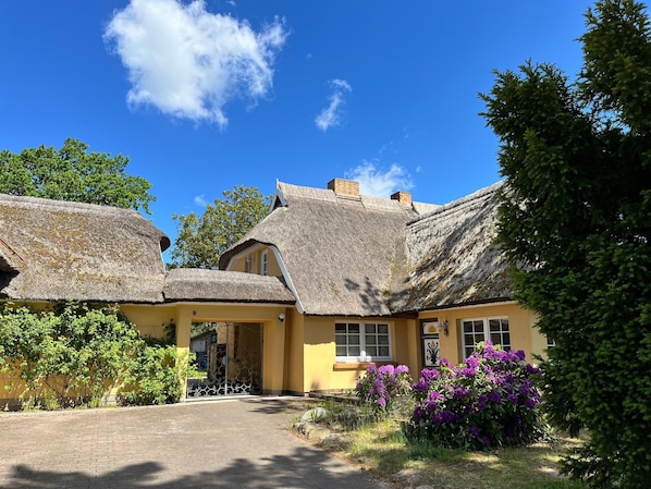 A traditional building with a thatched roof, a yellow facade, and a small garden with purple flowers.