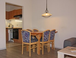 Un comedor con mesa y sillas de madera, una lámpara colgante y una cocina al fondo.