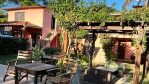 An outdoor patio with a wooden table and chairs, surrounded by greenery and a pergola. - Encanto dos Anjos (Búzios)