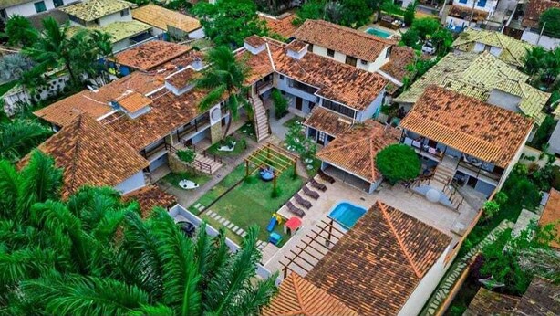 A residential area with multiple houses, each with a tiled roof, surrounded by greenery and a central courtyard with a swimming pool. - Encanto dos Anjos (Búzios)