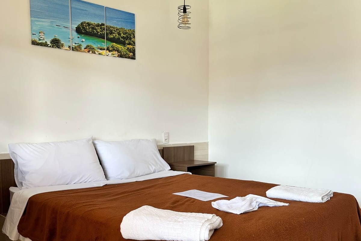 A neatly made bed with white pillows and a brown blanket, a painting of a coastal scene, and a simple hanging light fixture.