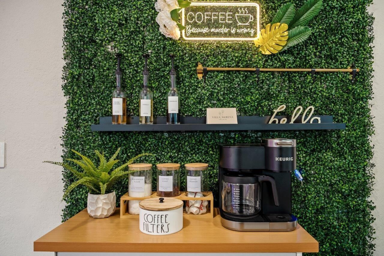A coffee station with a sign that reads "1 COFFEE Because murder is wrong." There are coffee filters, a coffee maker, and jars of coffee creamer on the counter.