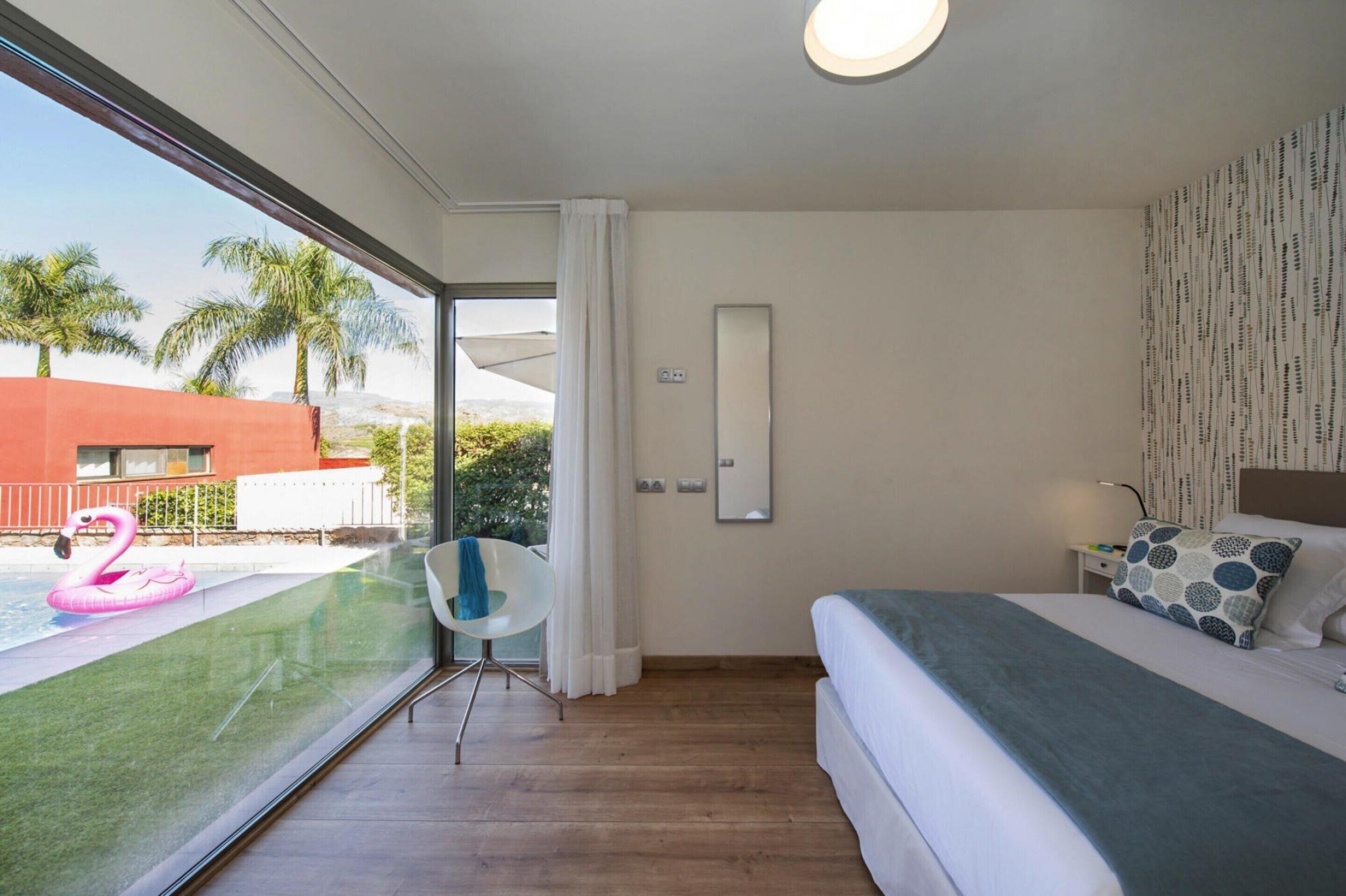 Een moderne hotelkamer met een groot raam, een bed, een stoel en uitzicht op een zwembad met een roze flamingo drijver.