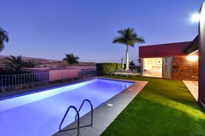 A swimming pool with a view of a desert landscape, a modern building with a stone facade, and a well-maintained lawn.
