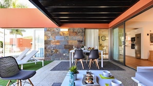 An outdoor dining area with a stone wall backdrop, a table set with dishes, and a modern kitchen visible through glass doors. - Holiday Home, Maspalomas (San Bartolomé de Tirajana)