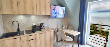 A modern kitchen with wooden cabinets, a black sink, and a dining area with a table and chairs. A television is mounted on the wall, and a balcony with a view of the sea is visible.