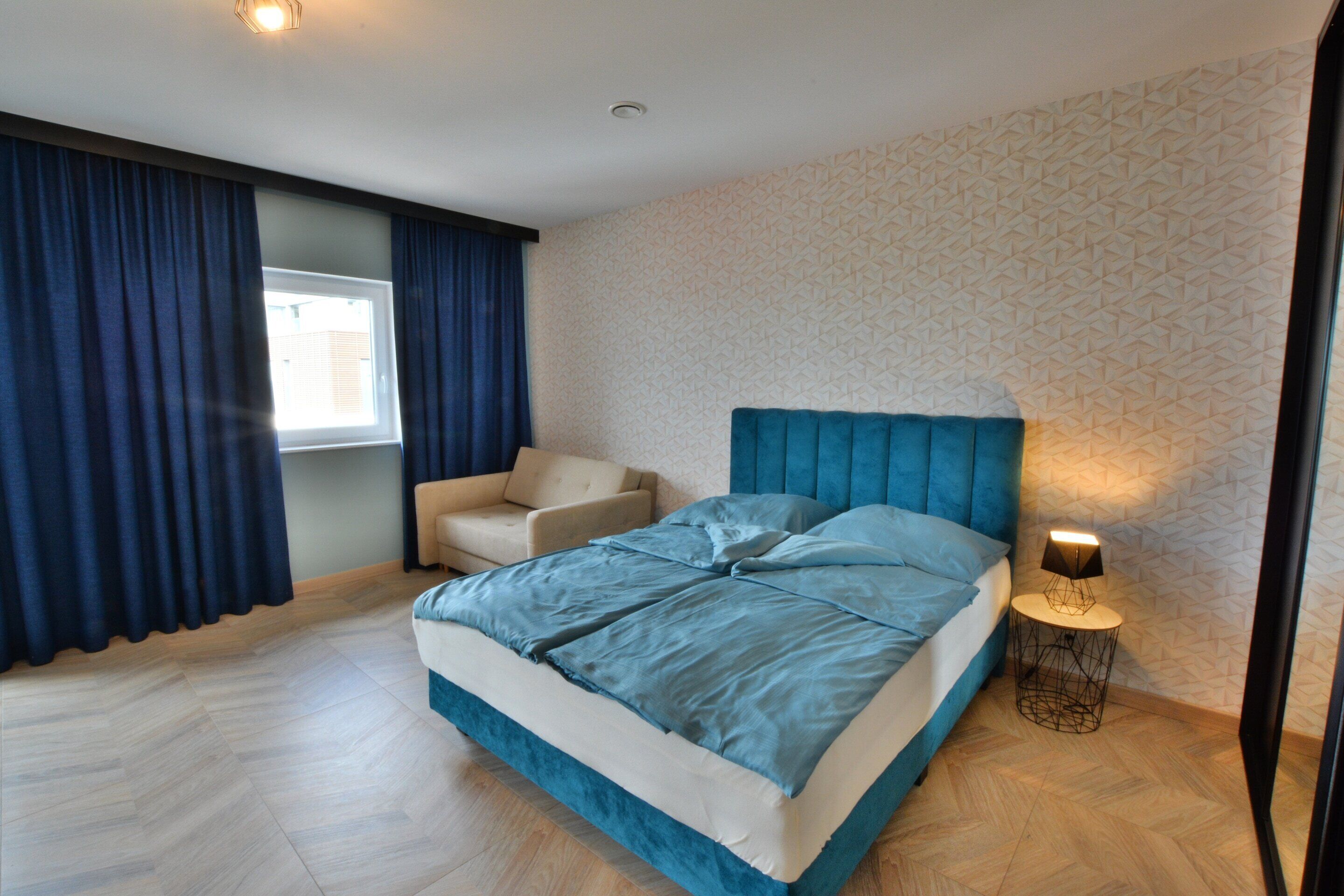 A bedroom with a bed, a chair, a bedside lamp, and a window with curtains.