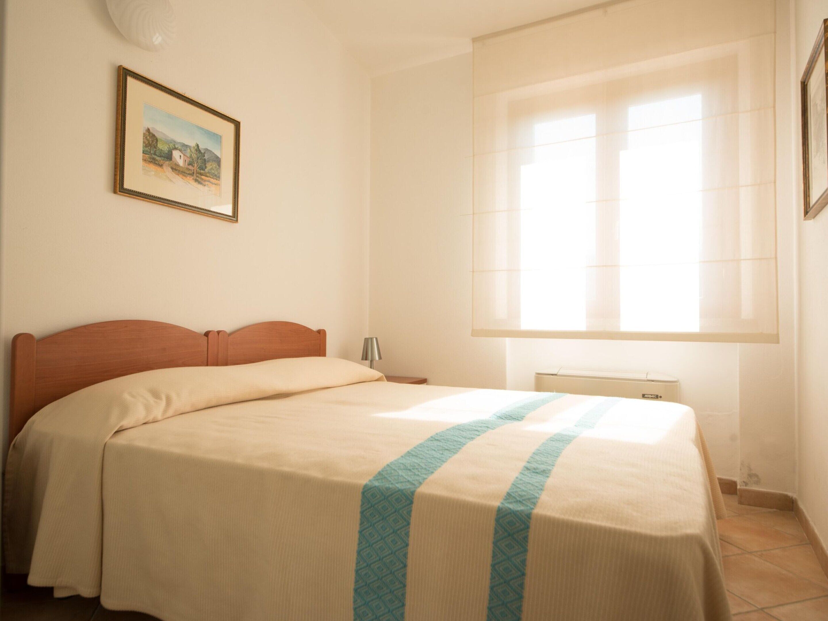 A bedroom with a bed, a nightstand, a lamp, a radiator, and a window with blinds.