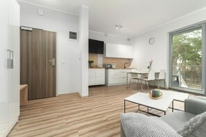 A modern apartment with a wooden floor, a kitchenette, a dining area, and a large window offering a view of the outdoors.