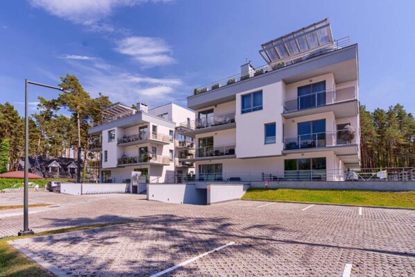A modern multi-story residential building with balconies, a paved driveway, and a clear sky.