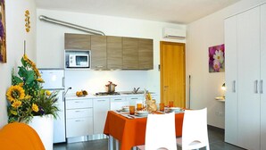 Une cuisine moderne avec des armoires blanches, un four à micro-ondes et une cuisinière. On y trouve une table à manger recouverte d’une nappe orange et entourée de chaises blanches. Un grand vase rempli de tournesols est placé sur la gauche.