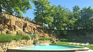 Una piscina con un muro de piedra y relieves, rodeada de césped y árboles.