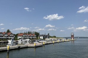 Moored boats line the dock, surrounded by buildings with red roofs and a distant lighthouse.