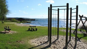 A playground with a swing set and climbing structure near a beach with a wooden building in the background. - Wunderschones Blockbohlenhaus am Plauer See (Plau am See)
