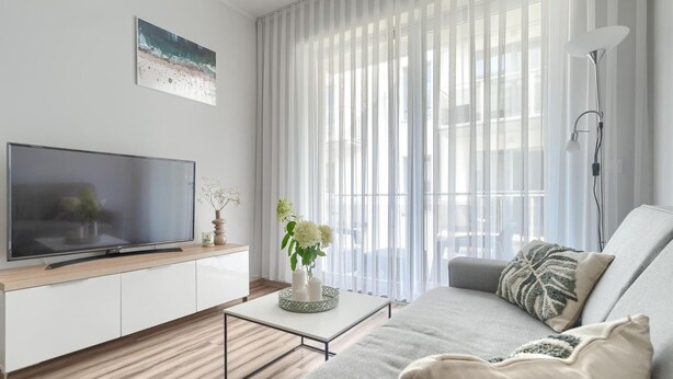 A modern living room with a flat-screen TV, a sofa, a coffee table, and a large window with sheer curtains. - Beautiful Apartment Very Close to the Beach (Rewal)