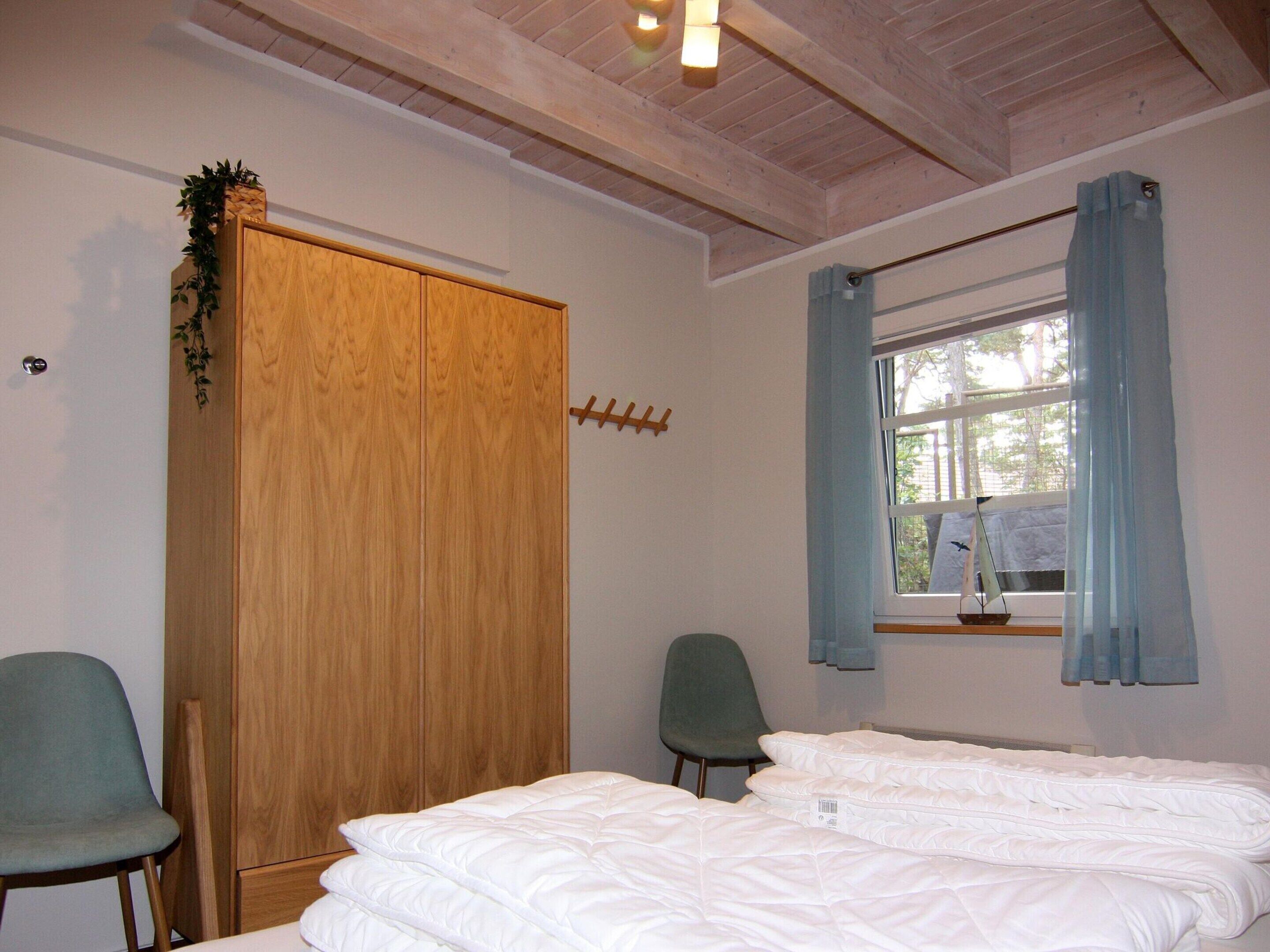 A bedroom with a wooden wardrobe, two green chairs, a bed with white bedding, a window with blue curtains, and a small plant on the wardrobe.