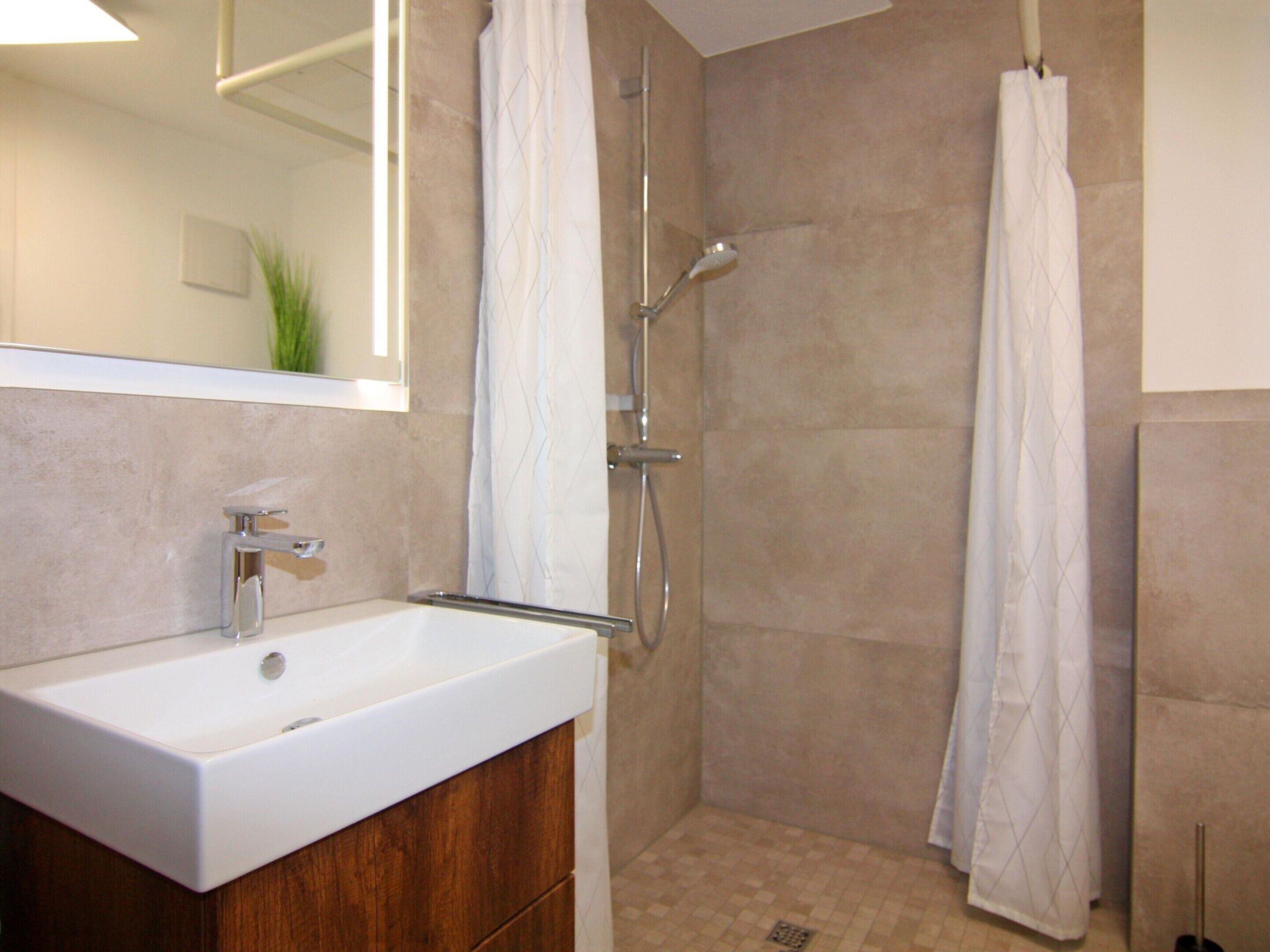 A modern bathroom with a white sink, wooden cabinet, and a walk-in shower with beige tiles.