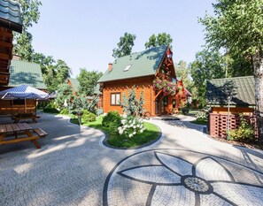 Una cabaña de madera con techo verde, rodeada por un jardín con flores y una zona circular empedrada.