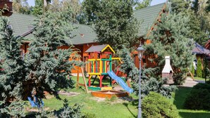 A playground with a slide and climbing structures, surrounded by trees and a building with a green roof. - Holiday Home Pobierowo Near Baltic Sea (Rewal)