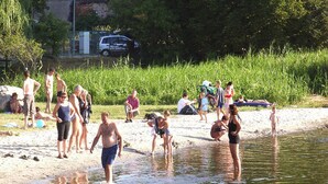 En gruppe mennesker samlet ved en sø med en sandstrand og en parkeret bil i baggrunden.