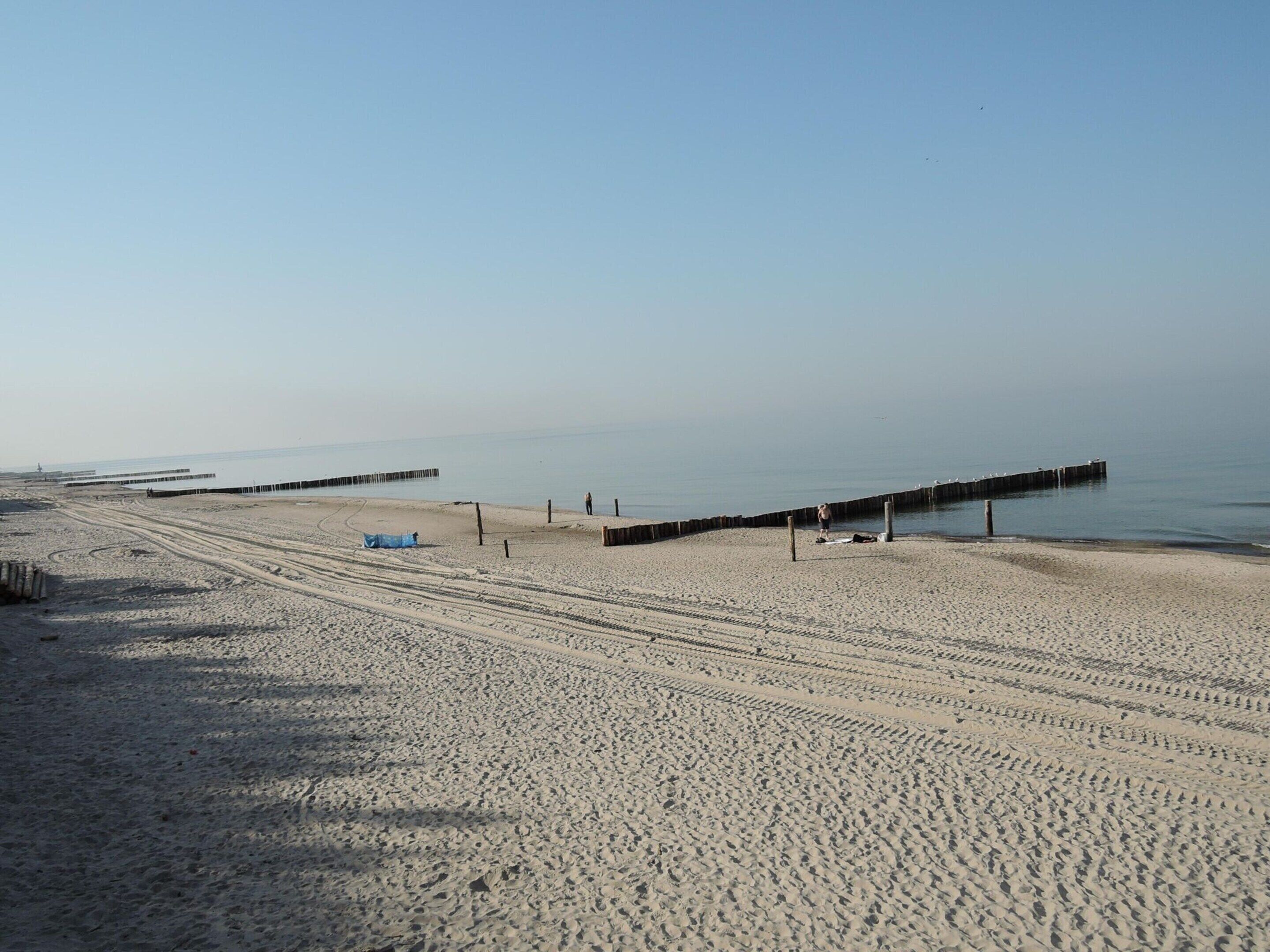 A wide sandy beach with a clear sky, a long pier extending into the water, and a few people walking along the shore.