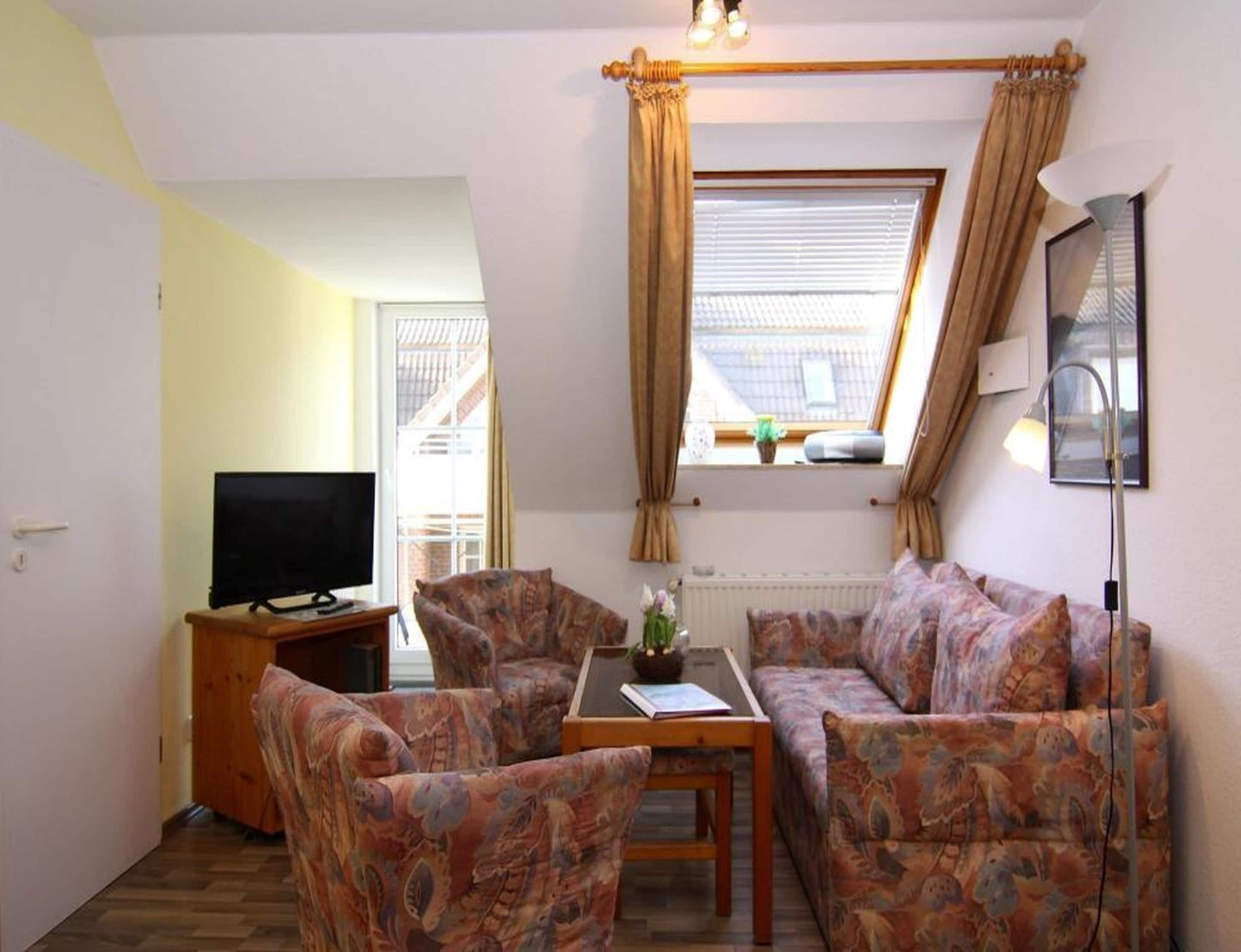 Una habitación con una televisión, dos sofás estampados, una mesita con una planta y una ventana con cortinas.