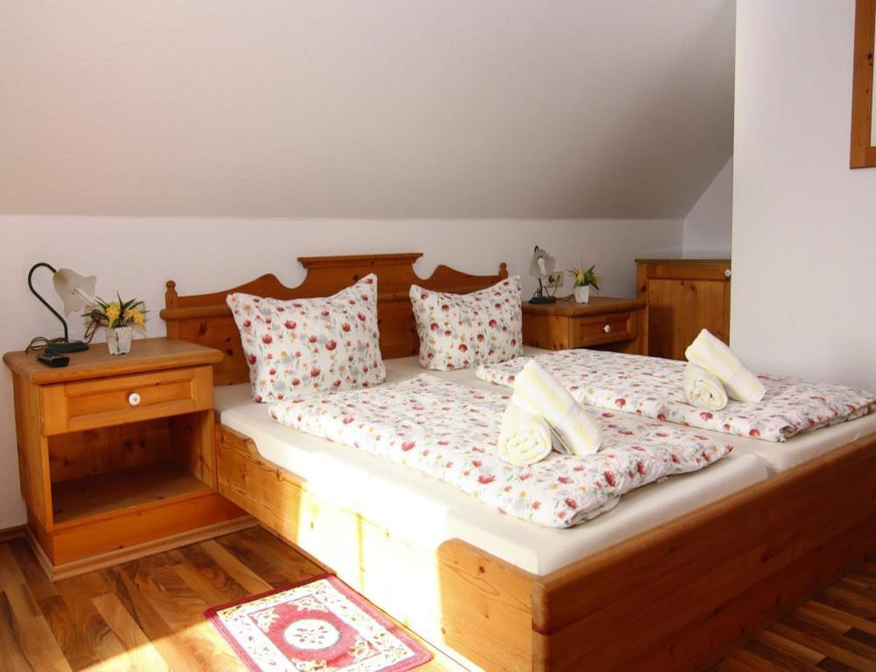 Una cama doble impecablemente arreglada con ropa de cama floral, cabecera de madera, mesitas de noche con lámparas y una pequeña planta en maceta.