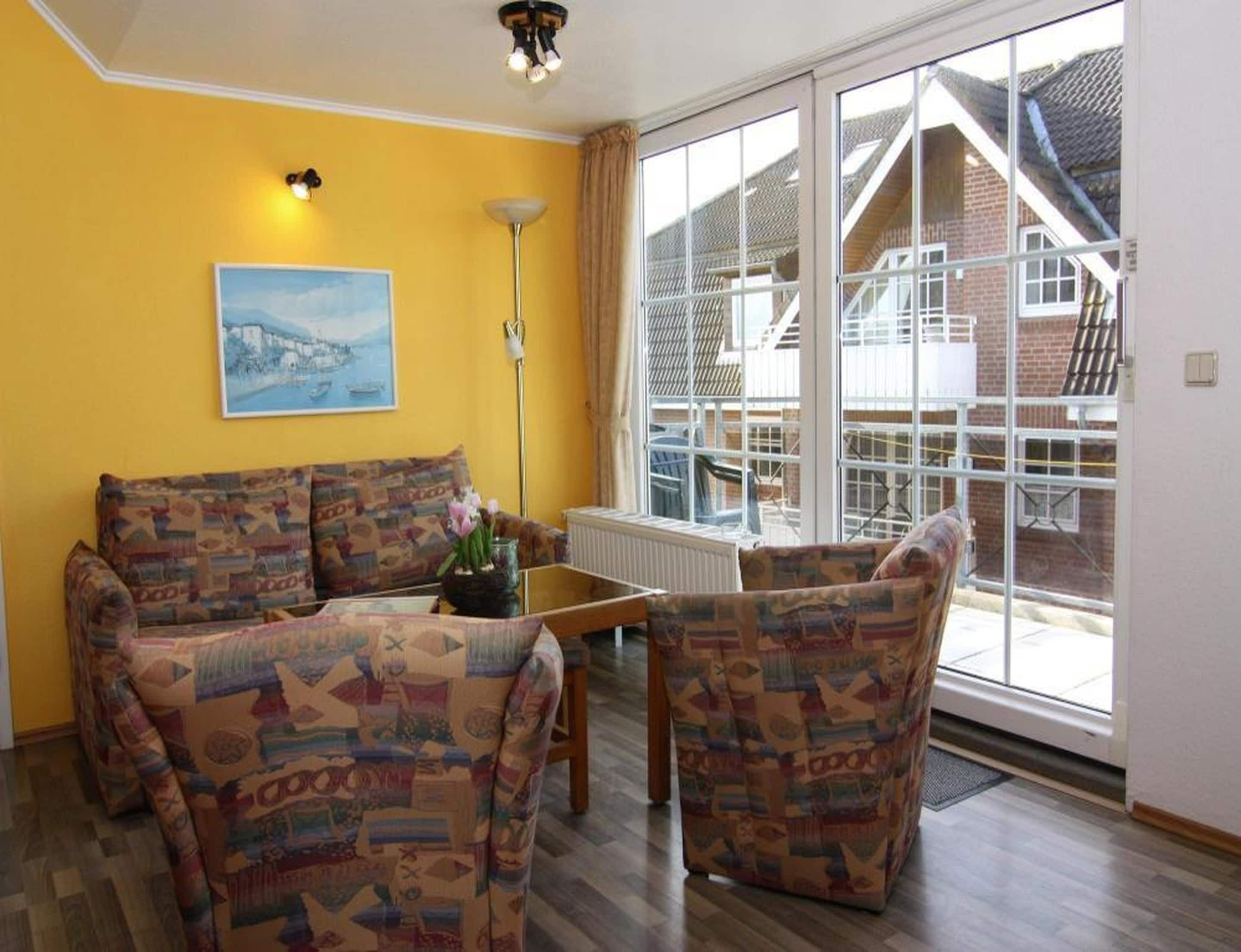 Un salón con sofás estampados, una mesa de centro, una lámpara de pie y un ventanal con vista a una casa.