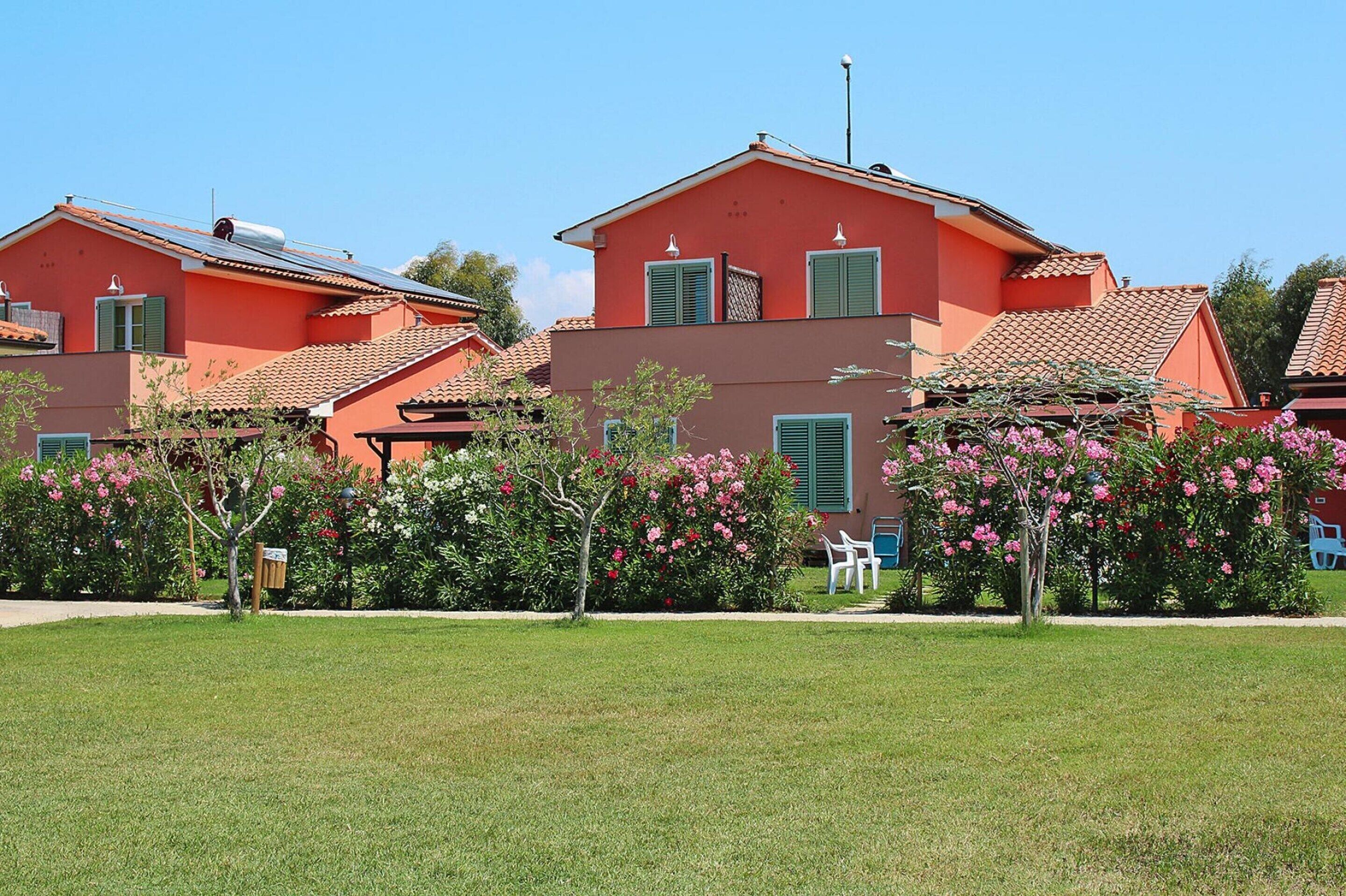 A row of red-roofed buildings with green shutters, surrounded by well-maintained gardens and pink flowering bushes.
