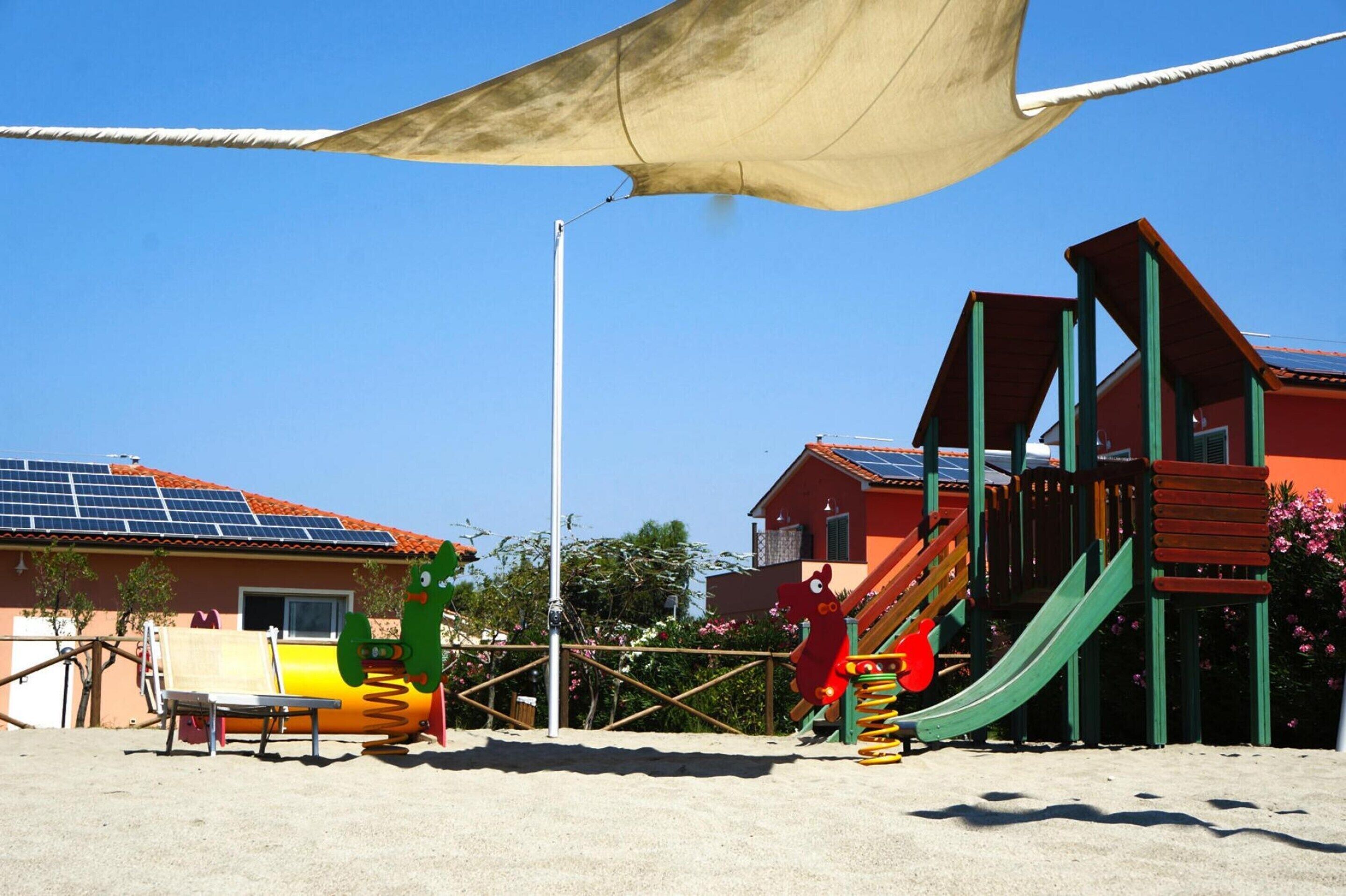 Ein Spielplatz mit Rutschen und Klettergerüsten, umgeben von Gebäuden mit Solaranlagen und blauem Himmel.