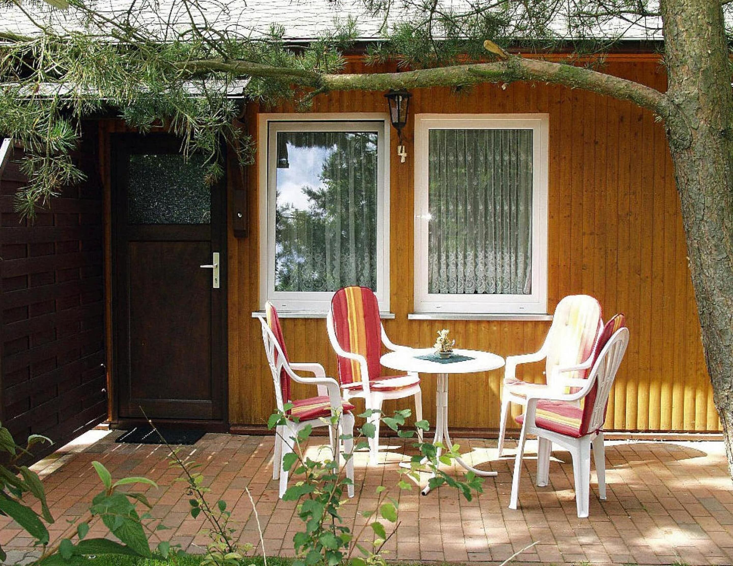 A wooden house with a brick exterior, two windows with curtains, and a small patio with white chairs and a table.