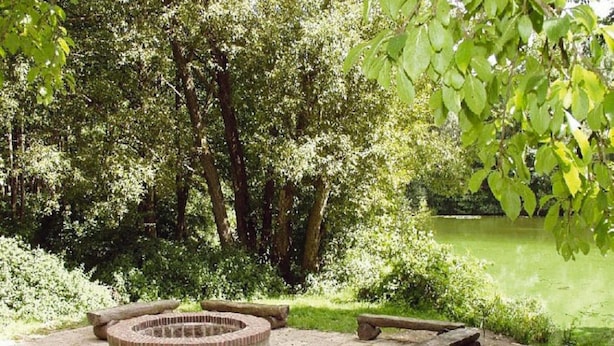 A circular stone fire pit surrounded by a paved area, with benches and a pond in a lush green park setting.