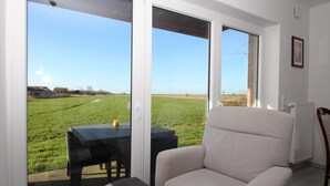 Una habitación con un gran ventanal corredero que da a un campo cubierto de césped, un sillón beige, una pequeña mesa con una silla y un radiador en la pared.