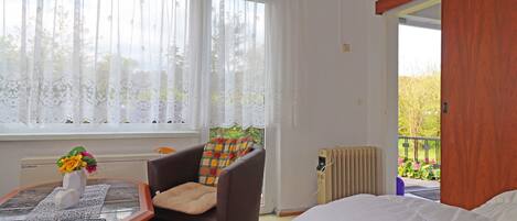 A hotel room with a bed, a chair with a cushion, a table with a vase of flowers, and a window with sheer curtains.